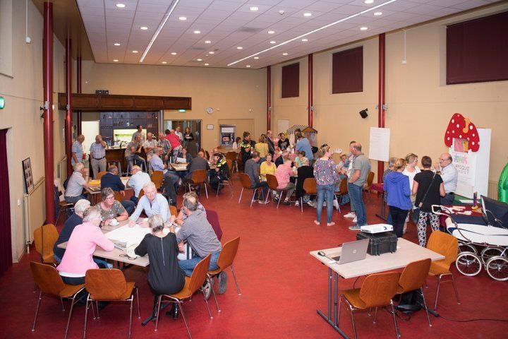 Mensen verzamelden zich in een grote zaal, zaten aan tafels en stonden, en namen deel aan een groepsactiviteit of vergadering.