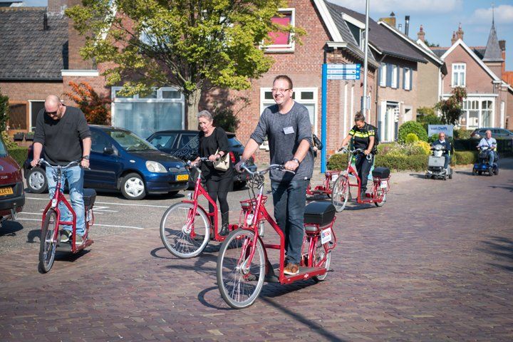 Mensen rijden op rode stepjes met zitjes in een zonnig, met bakstenen bestraat gebied, vlakbij geparkeerde auto's en gebouwen.