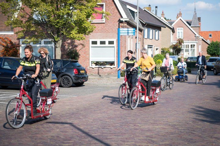 Politieagenten op rode fietsen patrouilleren door een bakstenen straat in een stad, waar ook inwoners op de fiets rondrijden.