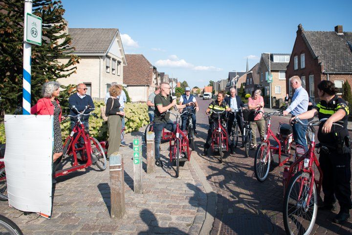 Groep op rode fietsen; straatbeeld met huizen. Mensen luisteren naar een man die spreekt. Een politieagente is aanwezig.
