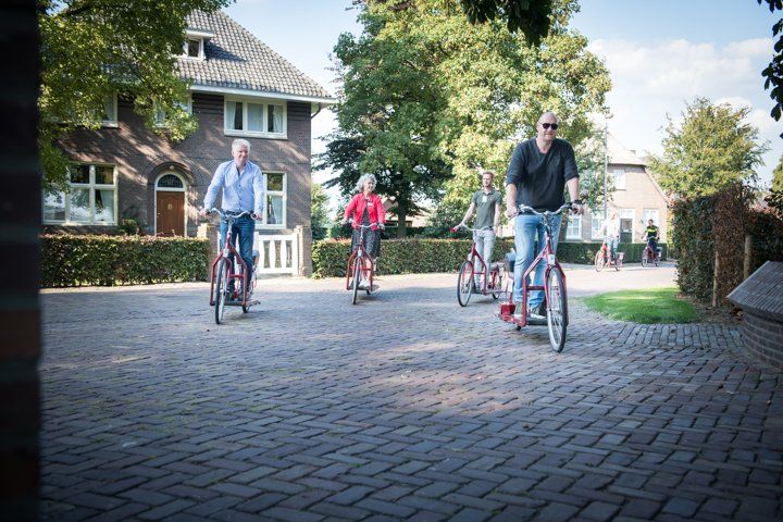 Vier mensen rijden op rode stepjes over een bakstenen pad, met bomen en huizen op de achtergrond.