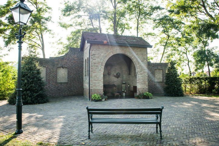 Bakstenen gebouw met boog en bank, onder bomen. Lantaarnpaal aan de linkerkant.
