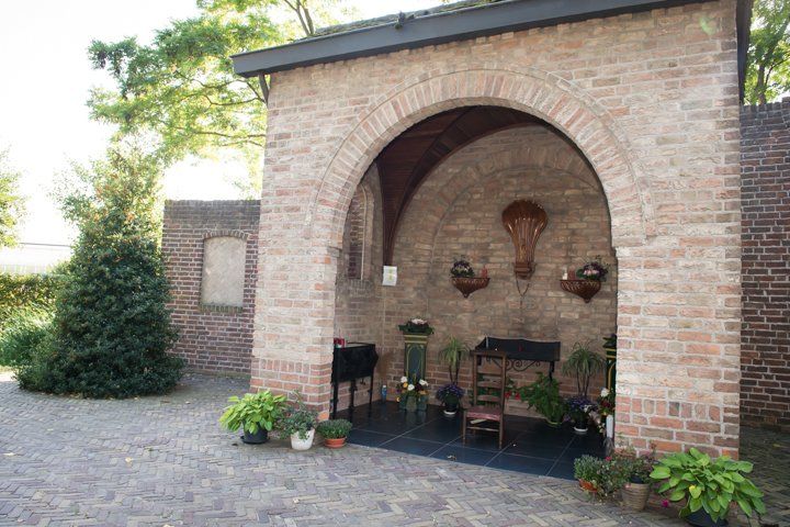 Een gewelfde nis van baksteen met een donkere tegelvloer, waarin tafels, stoelen en potplanten staan, en die aan de achterwand is versierd.