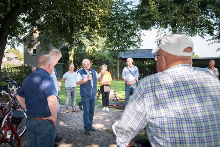 Een groep mensen luistert naar een man die buiten spreekt. Op de achtergrond zijn een verharde weg, fietsen, bomen en gebouwen te zien.