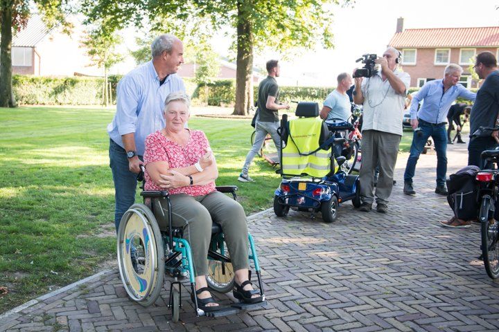 Een man duwt een vrouw in een rolstoel over een stenen pad; anderen zijn in de buurt. Een videocamera filmt hen.