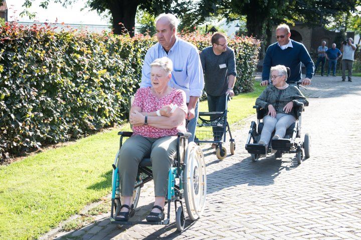 Mensen lopen en gebruiken mobiliteitshulpmiddelen op een zonnig pad met groen. Eén persoon in een rolstoel wordt voortgeduwd.