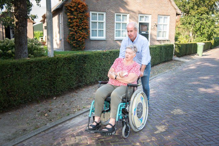 Een man duwt een persoon in een rolstoel over een stenen pad; op de achtergrond zijn een huis en heggen te zien.
