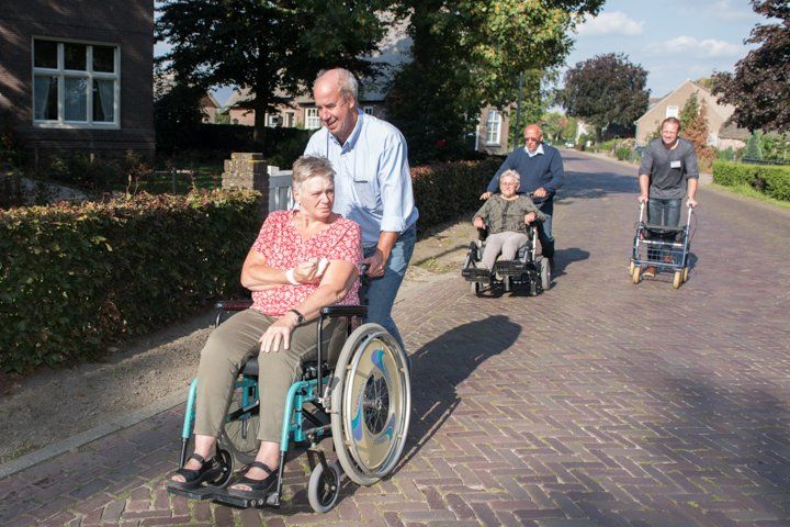 Een man duwt een persoon in een rolstoel over een stenen pad; anderen met mobiliteitshulpmiddelen volgen. Zonnige dag.