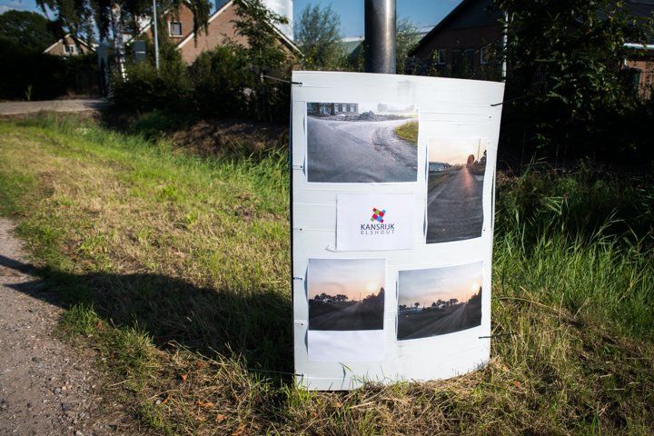 Poster met foto's op een paal in een grasveld, waarop een weg op verschillende tijdstippen van de dag te zien is.