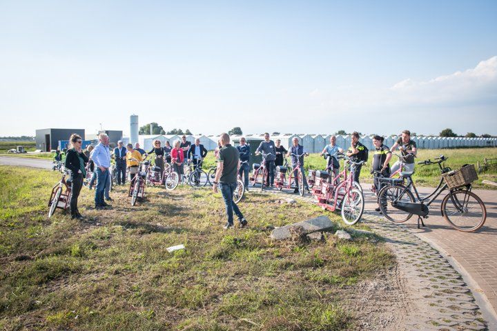 Een groep mensen met fietsen op een grasveldje vlakbij een weg en kassen op een zonnige dag.