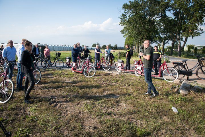 Mensen met fietsen verzamelen zich buiten; een man spreekt de groep toe op een zonnige dag.