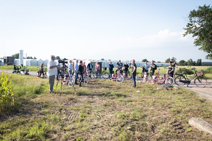 Een groep mensen op fietsen op een grasveld, waarschijnlijk voor een recreatieve fietstocht. Gebouwen in de verte.