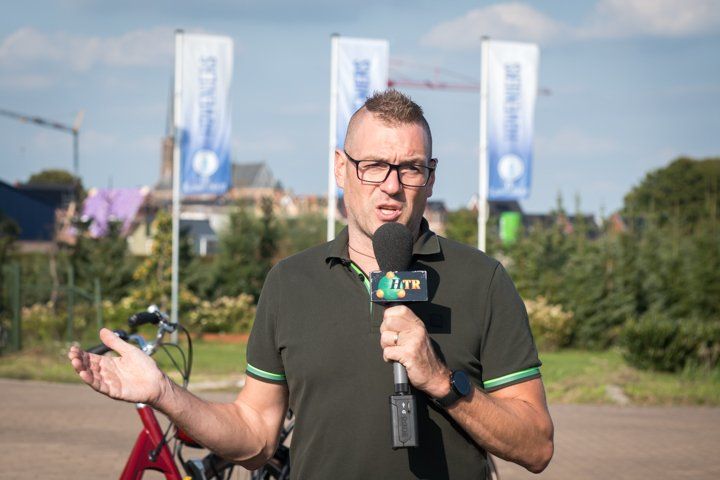Man met microfoon die buiten spreekt; gebouwen en vlaggen op de achtergrond.