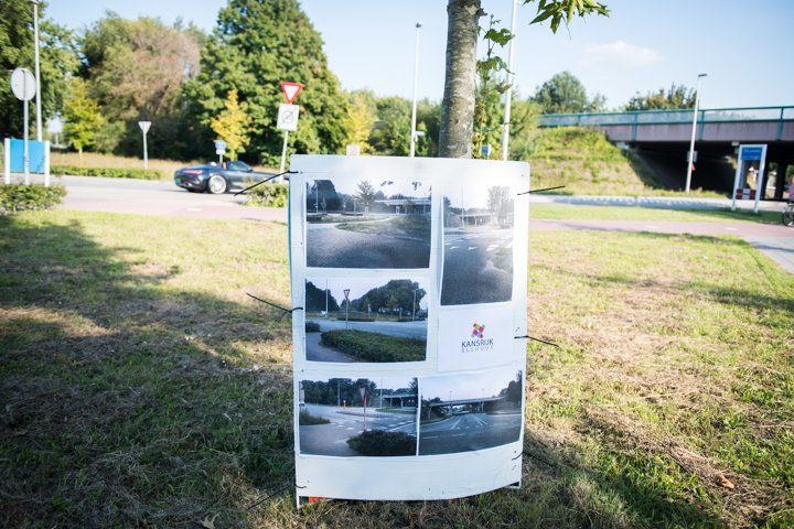 Foto's gemonteerd op een poster die tegen een boom leunt in een grasveld, met een straat en een brug op de achtergrond.