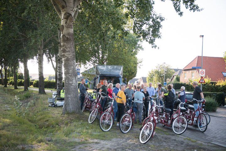 Een groep mensen met aangepaste fietsen verzamelde zich bij een weg en een geparkeerde bestelwagen, omringd door bomen en huizen.