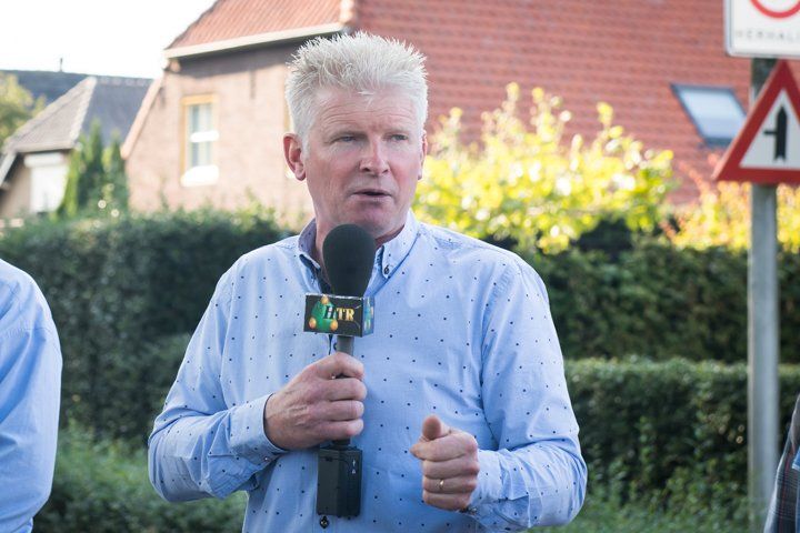 Een man in een blauw shirt met een microfoon in zijn hand staat buiten, vlakbij een weg met begroeiing en een bord.