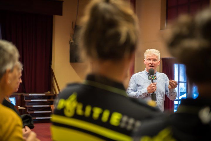 Een man spreekt in een microfoon tot een groep, waarschijnlijk in een zaal. Sommigen dragen politie-uniformen.