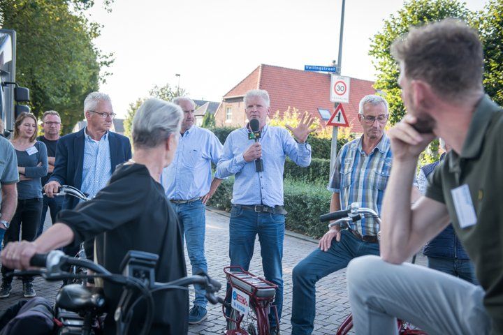 Een man met een microfoon gebaart naar een groep mensen en fietsen buiten op straat.