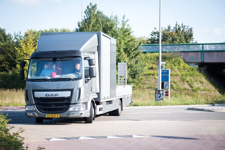 Een zilverkleurige DAF-vrachtwagen rijdt over een weg en passeert een blauw tolhuisje onder een brug; twee personen zijn zichtbaar in de cabine.