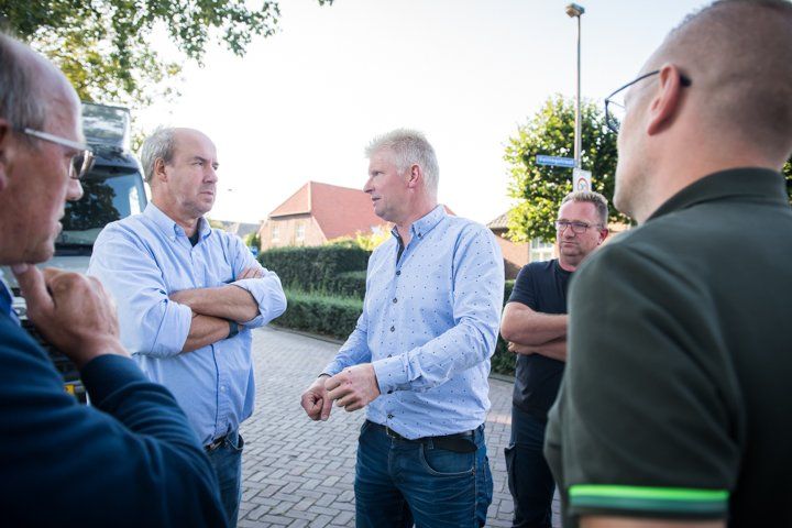 Een groep mannen staat op straat te praten. Sommigen hebben hun armen over elkaar geslagen en kijken serieus. Op de achtergrond zijn een bakstenen pad, struiken en gebouwen te zien.