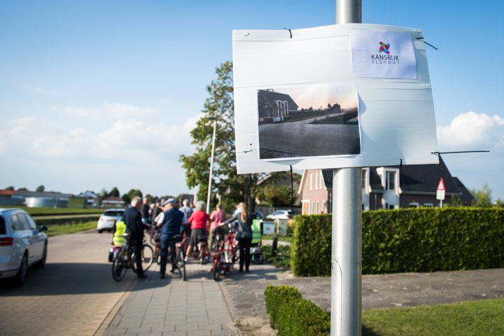 Een groep fietsers bij een bord met een foto van een waterweg en een logo op een zonnige dag.