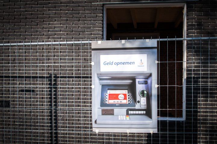 De geldautomaat is beveiligd achter een gaashekwerk tegen een bakstenen muur. Boven de geldautomaat bevindt zich een raam.