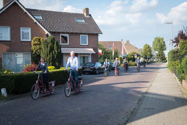 Mensen rijden op grote rode scooters door een woonstraat met bakstenen huizen onder een blauwe hemel.