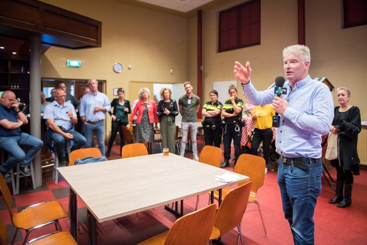 Een man spreekt in een microfoon en gebaart naar het publiek in een zaal.