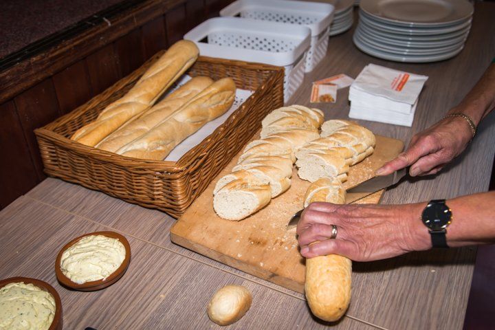 Iemand snijdt een stokbrood op een houten plank; een broodmandje, boter, borden en servetten staan ​​in de buurt.