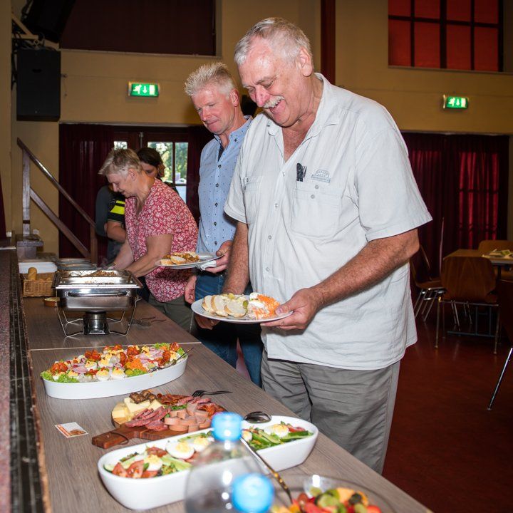 Mensen bij een buffet, die hun eten uitkiezen. Binnen, houten tafels, een man glimlacht terwijl hij een bord vasthoudt.