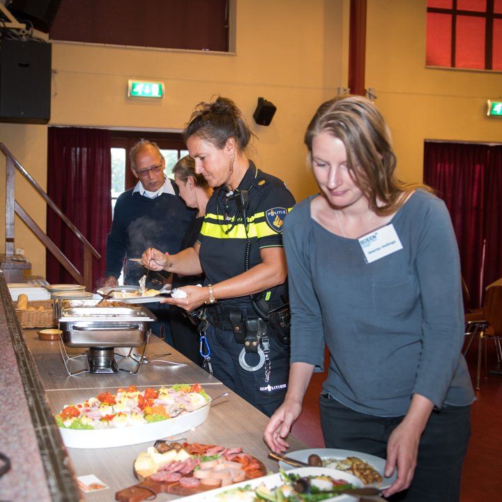 Mensen bij een buffet. Een agent in uniform serveert eten samen met anderen in een felverlichte zaal.