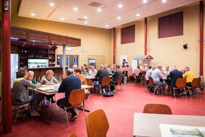Mensen zitten aan tafels in een gemeenschapshuis met een bar en een kleine kraam.