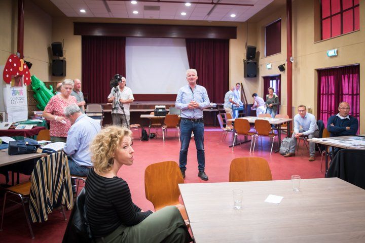 Een man spreekt in een gemeenschapszaal, met mensen die zitten, tafels en een projectiescherm op de achtergrond.