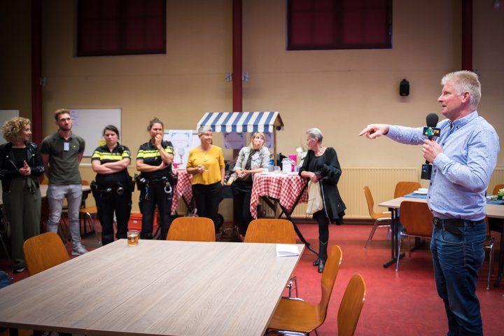 Een man met een microfoon wijst naar een groep mensen die in een kamer staan. Sommigen dragen uniformen.