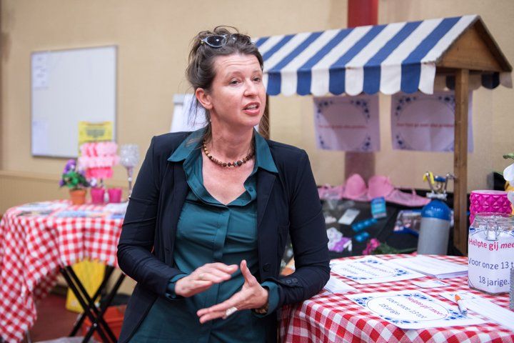 Een vrouw in een blazer spreekt en gebaart bij een kleine marktkraam met een geruit tafelkleed en een blauw-wit afdak.
