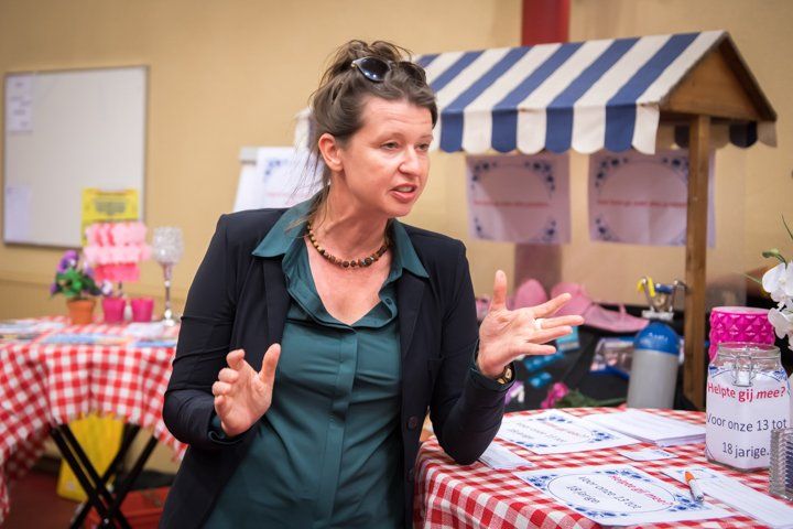 Vrouw in groene blouse gebaart bij een tafel met een rood-wit geblokt tafelkleed, met daarachter een kleine kraam met luifel.