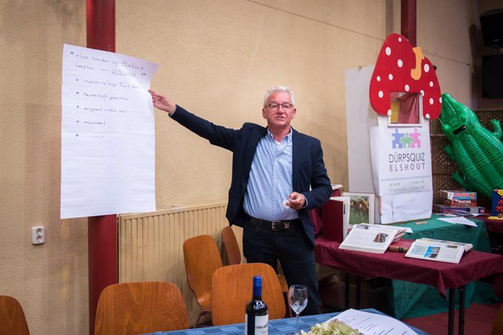 Een man geeft een presentatie aan een tafel met een poster. Een paddenstoeldecoratie en papieren liggen ernaast.