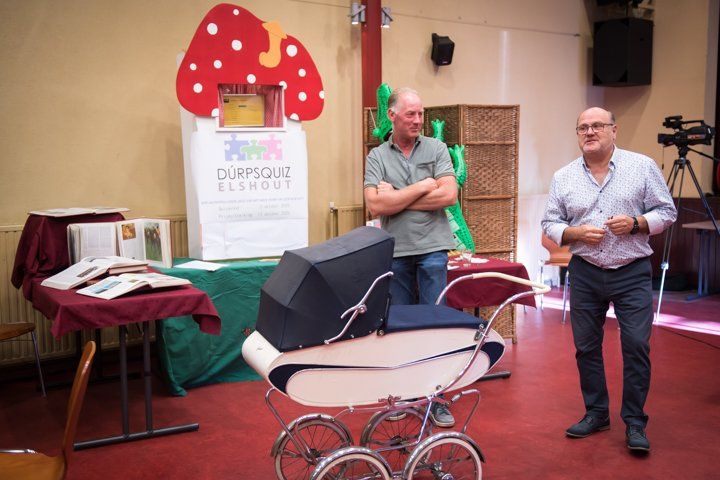 Twee mannen staan ​​bij een antieke kinderwagen in een kamer met een paddenstoeldecoratie en een tafel.