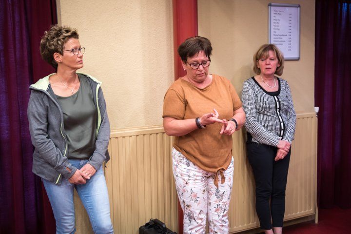 Drie vrouwen staan; een gebaart, twee kijken toe. Een kamer met gordijnen, een deur en een lijst aan de muur.