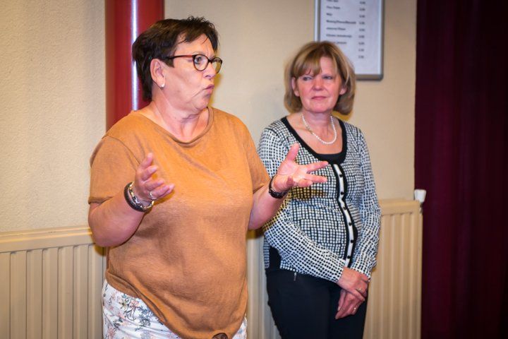 Twee vrouwen in een kamer, de ene gebaart terwijl ze spreekt, de andere staat naast haar.