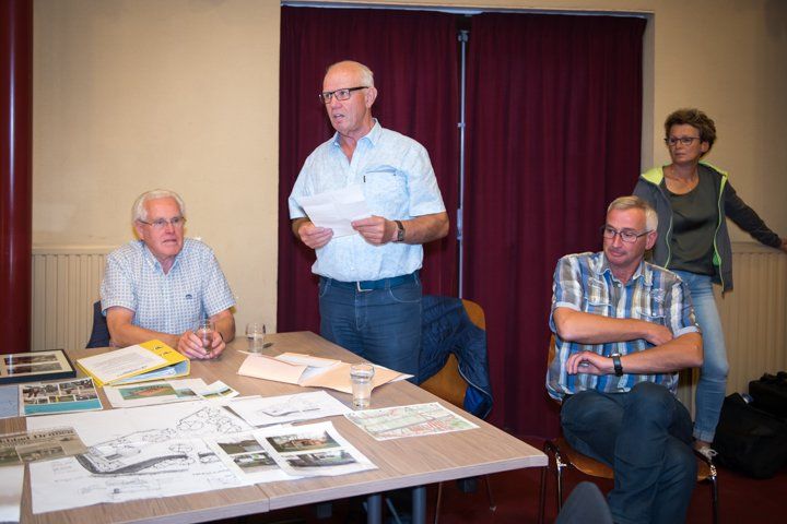 Vier mensen zitten aan een tafel; een man leest voor uit papieren terwijl anderen luisteren, in een kamer.