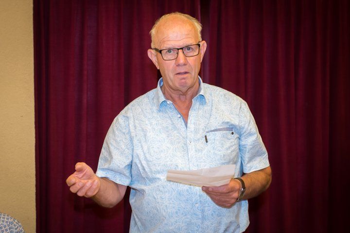 Een man in een lichtblauw shirt spreekt, houdt papieren vast en kijkt bezorgd; op de achtergrond bevindt zich een rood gordijn.
