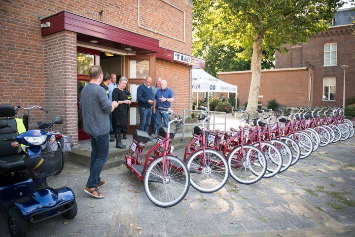 Een groep mensen staat buiten een bakstenen gebouw met een rij rode tandemfietsen. Op de voorgrond is een scooter te zien.
