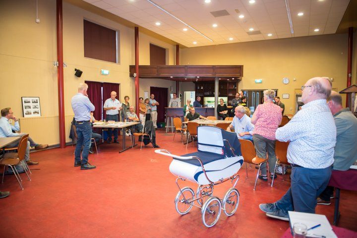 Mensen hadden zich verzameld in een kamer met een rode vloer en tafels, sommigen stonden bij een ouderwetse kinderwagen, wellicht voor een vergadering.