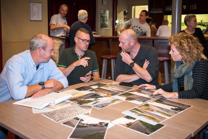 Een groep mensen bekijkt en bespreekt foto's aan een tafel. Op de achtergrond staan ​​anderen aan een bar.