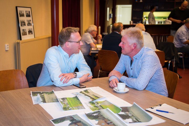 Twee mannen in lichtblauwe overhemden zitten aan een tafel en bekijken foto's; op de achtergrond zijn meerdere mensen te zien.