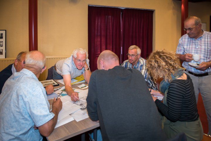 Een groep mensen zit aan een tafel en bekijkt documenten. Sommigen leunen voorover en voeren een gesprek. Een man houdt een koffiekopje in de buurt.