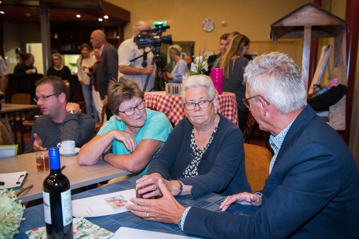 Een man in pak spreekt met twee vrouwen aan een tafel. Anderen kijken toe in een kamer.