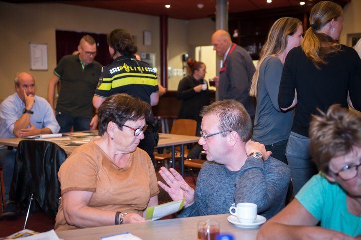 Mensen praten aan tafels in een kamer, met een politieagent op de achtergrond.