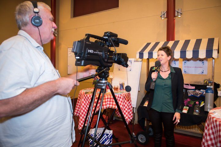 Een vrouw spreekt in een microfoon terwijl een cameraman haar filmt voor een kleine marktkraam.